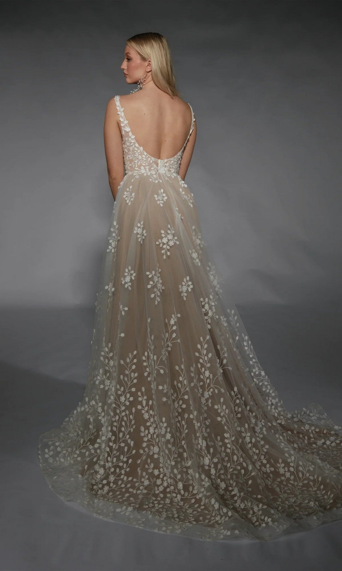 A woman wears the Long White Dress: Alyce 7132, a floor-length ivory bridal gown with a sleeveless design, deep V-back, and intricate 3D floral appliqué embroidery, standing against a gray background.